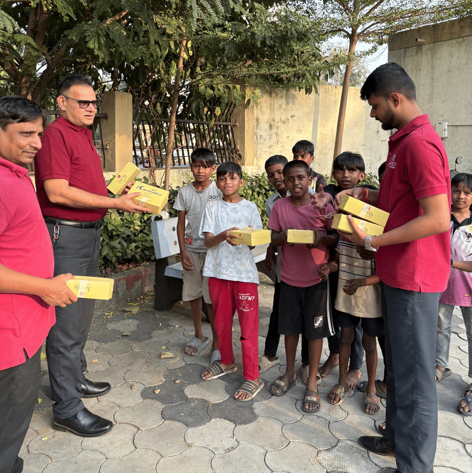 Group of men in red shirts distributing food boxes to smiling children outdoors near trees and a wall, during a community outreach activity.