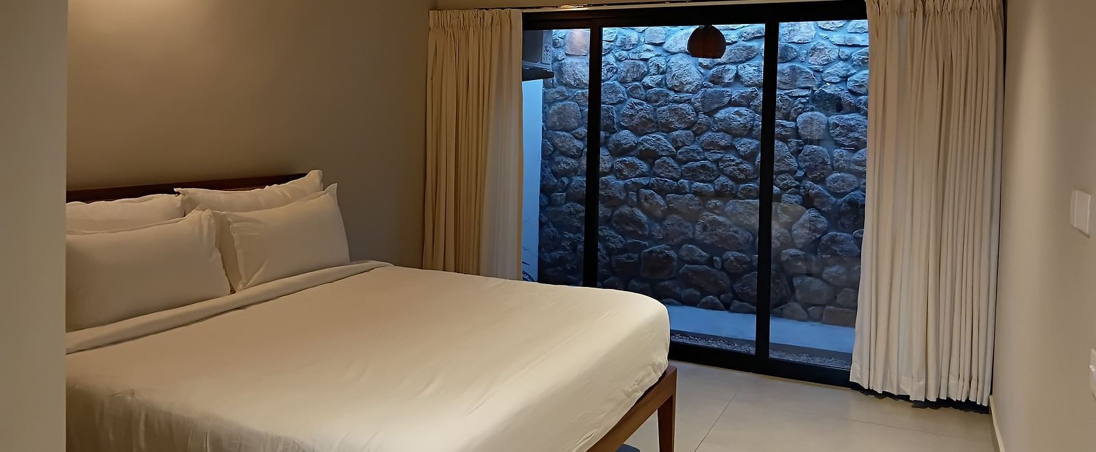 A cozy bedroom at Vaasta Vagamon with a neatly made bed, a ceiling fan, and a large window overlooking a stone wall.