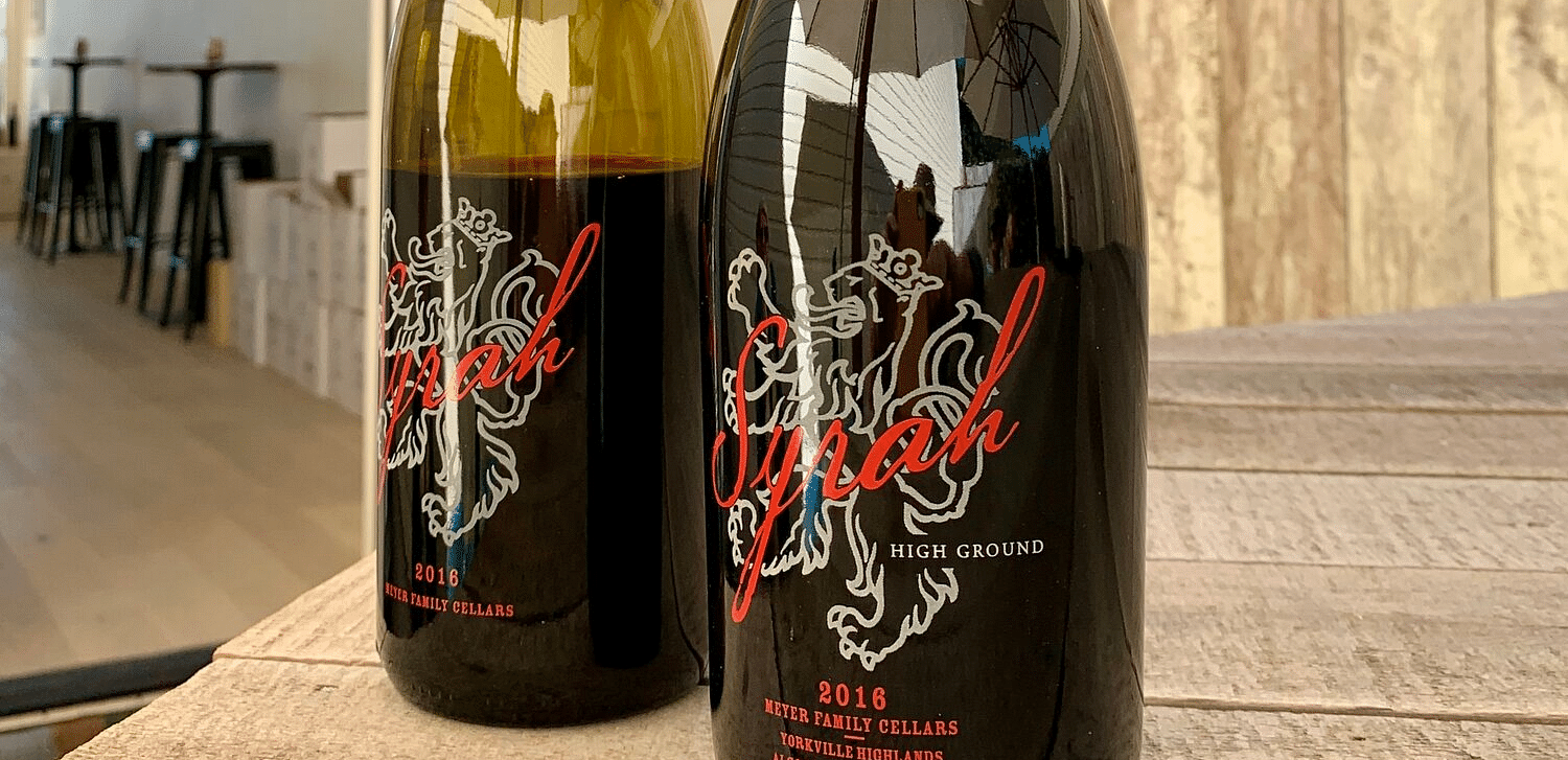 Two bottles of wine, one labeled High Ground, sit on a wooden table in front of a building.