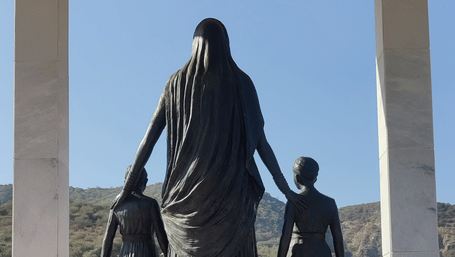 Panna Dhai statue holding the hands of two children, standing beneath a marble pavilion with hills and a clear sky in the background.