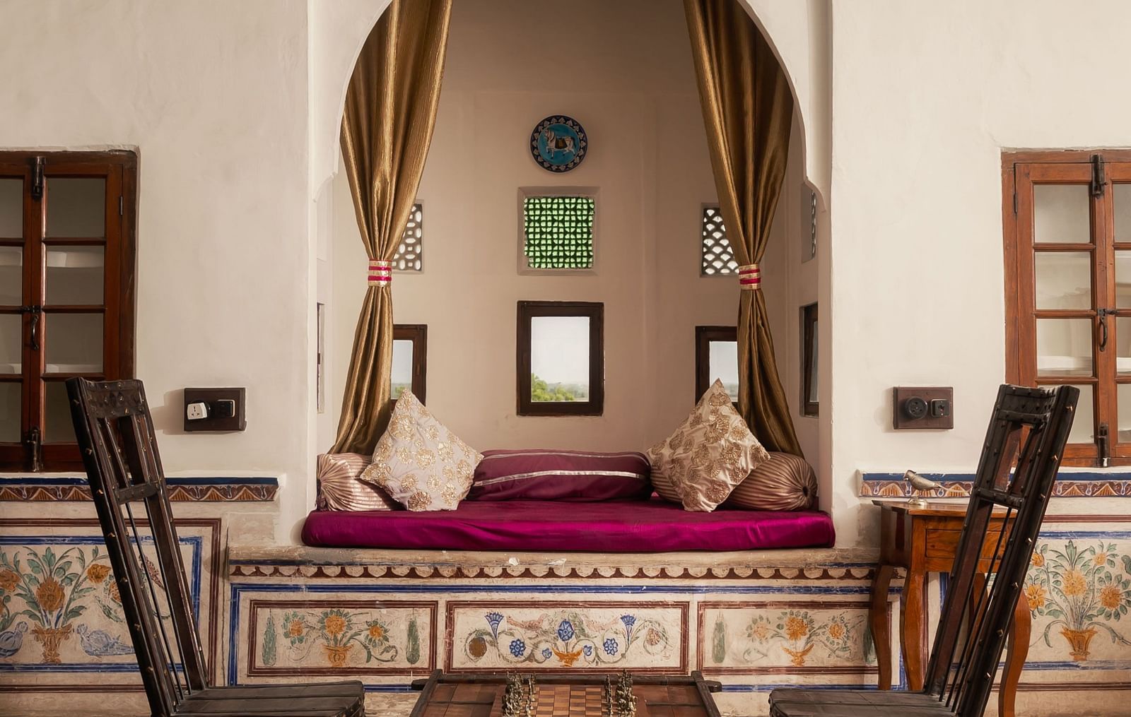 A stylish seating area at Fort Barli, featuring tall chairs, a low table, and an alcove with plush cushions, is illuminated by a striking blue chandelier.