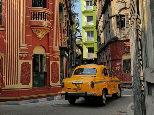 Iconic Kolkata yellow taxi passing through a historic neighbourhood with colourful colonial architecture.