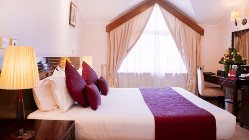 Standard Room featuring a plush bed by a window with curtains on  and a table lamp on the nightstand and a desk and a chair in the distance next to the window