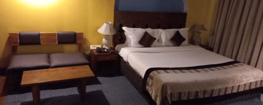Guest room at The Bristol Hotel, Gurgaon with a king-size bed, an arched headboard, striped bedspread, brown cushions, a bedside lamp, and a cushioned bench.