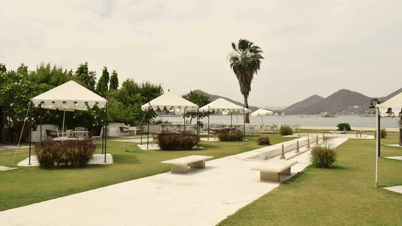 Outdoor seating area with white tents, benches, and lush green lawn by a lake with hills and Fateh Sagar Lake in the background at Ram Pratap Palace, Udaipur.