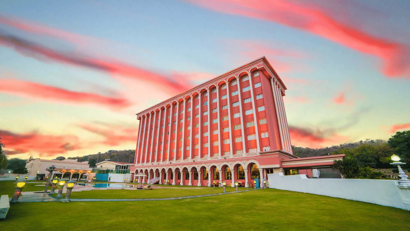 Modern multi-storey hotel building with arched ground-floor walkways, landscaped lawns, and a vibrant sunset sky in the background.