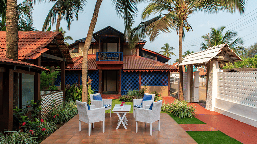 Outdoor seating area with white chairs and tables surrounded by palm trees, greenery and red-roofed buildings.