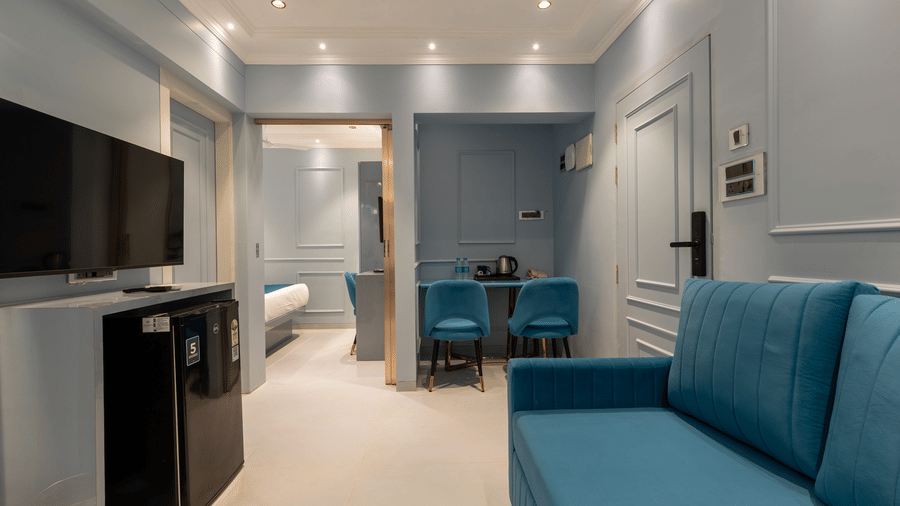 A modern living room featuring a plush blue sofa, flat-screen TV, and an open doorway leading to a bedroom - Home2 Residency, Mumbai Airport