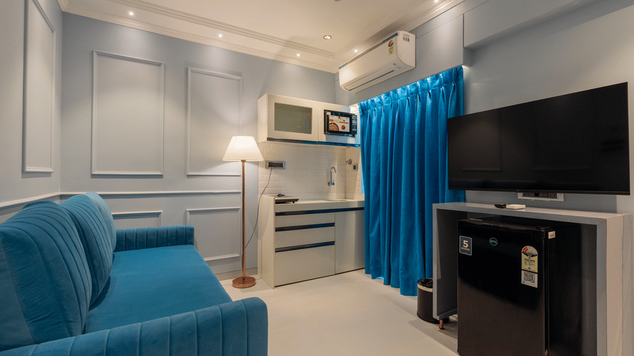 A studio living space with a blue sofa, wall-mounted television, and a compact kitchenette - Home2 Residency, Mumbai Airport