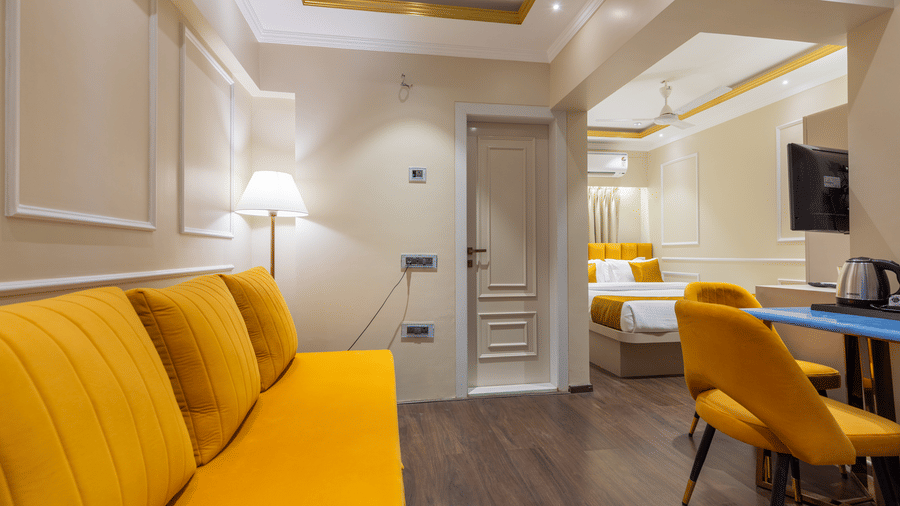A vibrant studio living space with a yellow sofa, dark wood flooring, and a bright bedroom visible in the background - Home2 Residency, Mumbai Airport