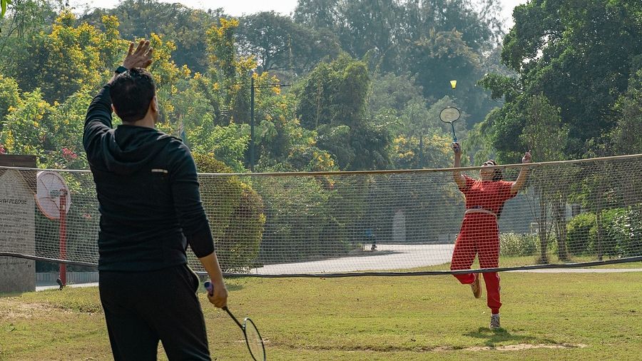 Two people playing badminton on a grassy outdoor court surrounded by trees, one mid-jump while hitting the shuttle with a racquet | Karma Chalets