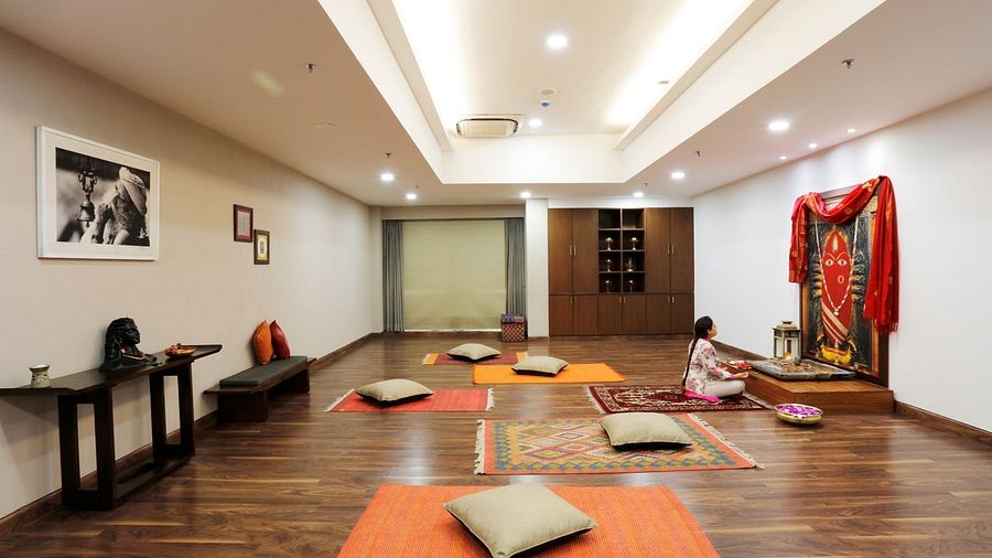 Yoga room with wooden flooring, orange mats, floor cushions, framed photos on the wall, and a red decorative cloth on a stand.