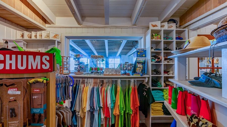 Clothing shop featuring casual wear, a branded display, and shelves stocked with shirts, shorts, and other beach gear.