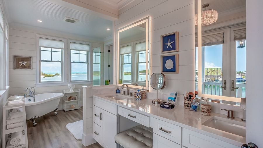 a bright and airy bathroom with a bathtub, shelves with towels, a makeup area with two huge mirrors, drawers and a chair with windows that overlooks the sea at Abaco Inn.