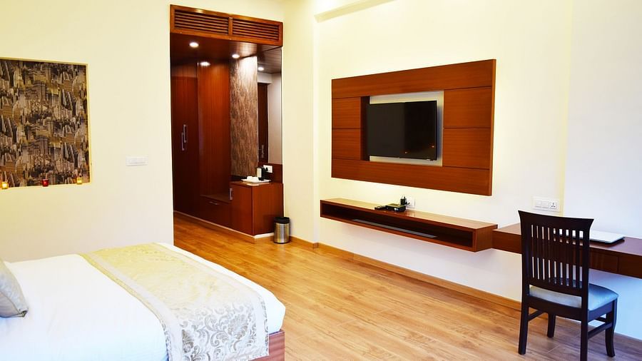 The image shows a hotel room with a large bed, a TV mounted on the wall, and a desk with a chair  | DLS Serenity Resort and Spa