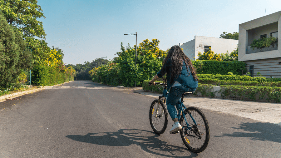 A girl riding a bicycle on a paved road with houses and green trees on either side, captured from behind in bright daylight | Karma Chalets