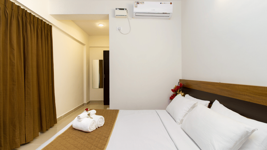 Hotel room with a double bed, brown curtains, an air conditioner, and a folded towel arrangement at Sanctum Suites Whitefield, Bangalore.
