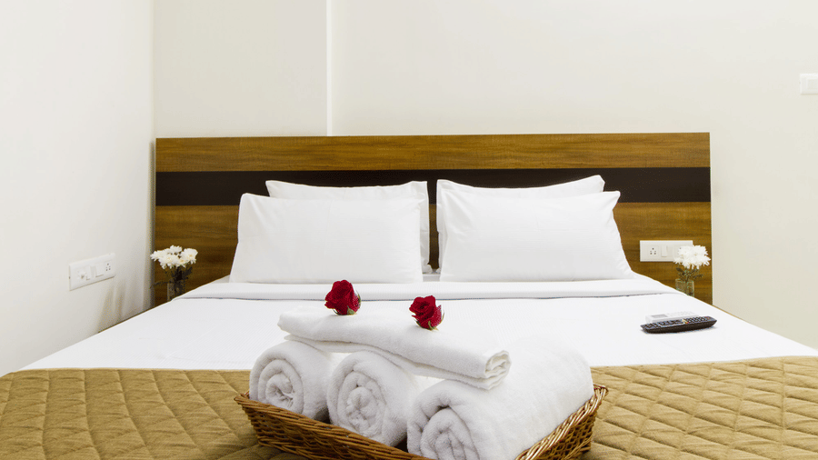 Close-up of a hotel room bed with white linen, a rose, and neatly folded towels at Sanctum Suites Whitefield, Bangalore.