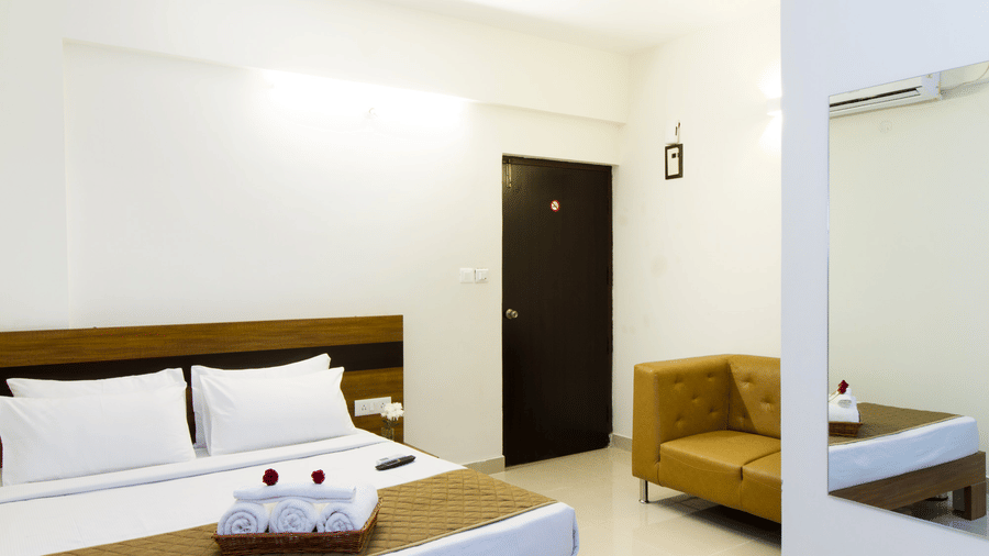 Airy hotel room with a double bed, a sofa, and a wooden door at Sanctum Suites Whitefield, Bangalore.