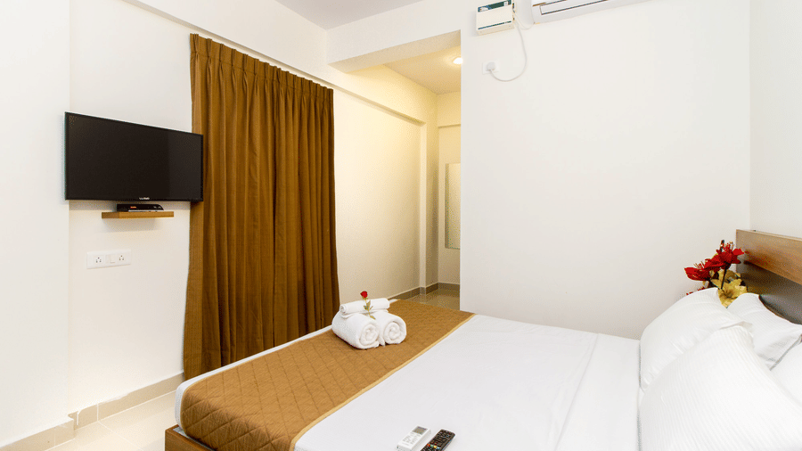 Hotel room with a double bed, a wall-mounted television, brown curtains, and an air conditioner at Sanctum Suites Whitefield, Bangalore.