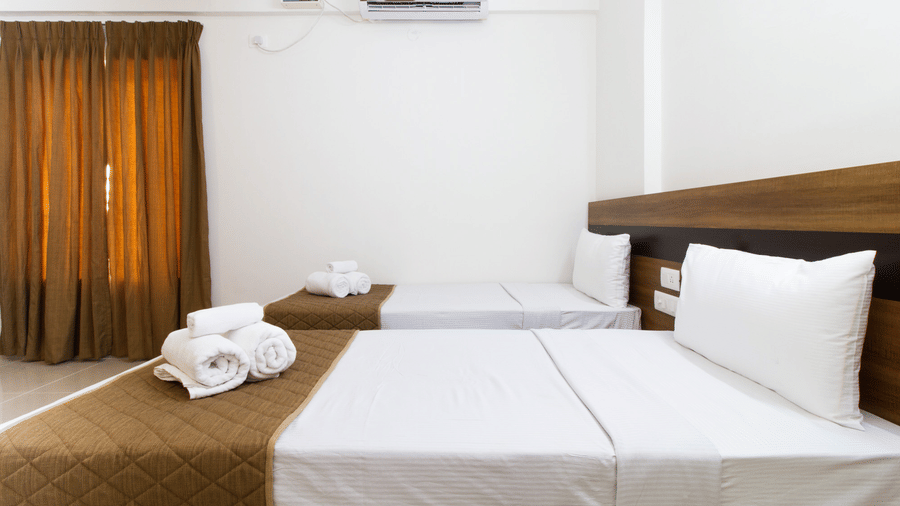 Hotel room with a double bed, white linen, brown curtains, and an air-conditioner at Sanctum Suites Whitefield, Bangalore.