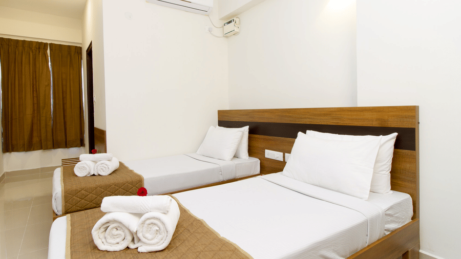 Hotel room with a double bed, brown curtains, an air conditioner, and a neatly arranged towel set at Sanctum Suites Whitefield, Bangalore.