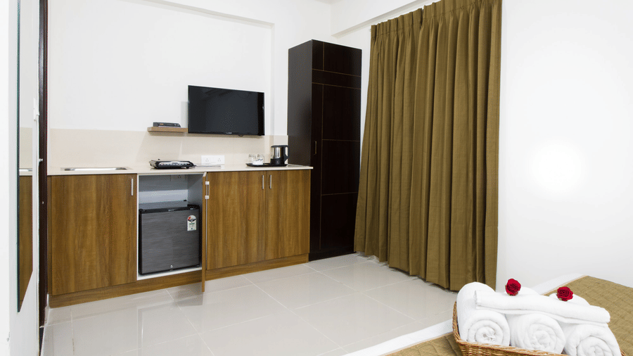 Hotel room with a television, minibar, wooden cabinetry, and a bed with a decorative towel arrangement at Sanctum Suites Whitefield, Bangalore.