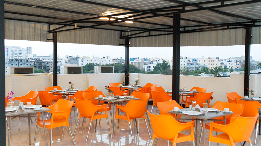 Rooftop dining area with orange chairs, round tables, and a pergola structure at Sanctum Suites Whitefield, Bangalore.