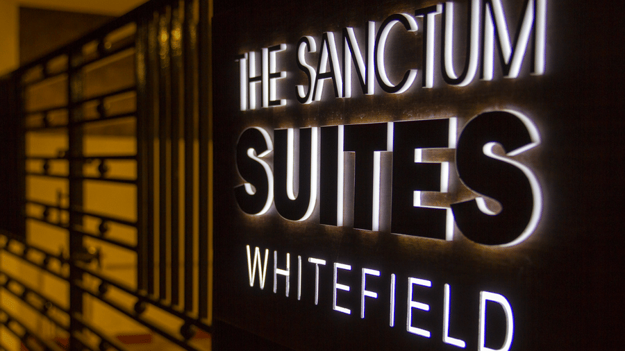 Illuminated backlit brand signage of The Sanctum Suites Whitefield against a dark wooden panel at Sanctum Suites Whitefield, Bangalore.