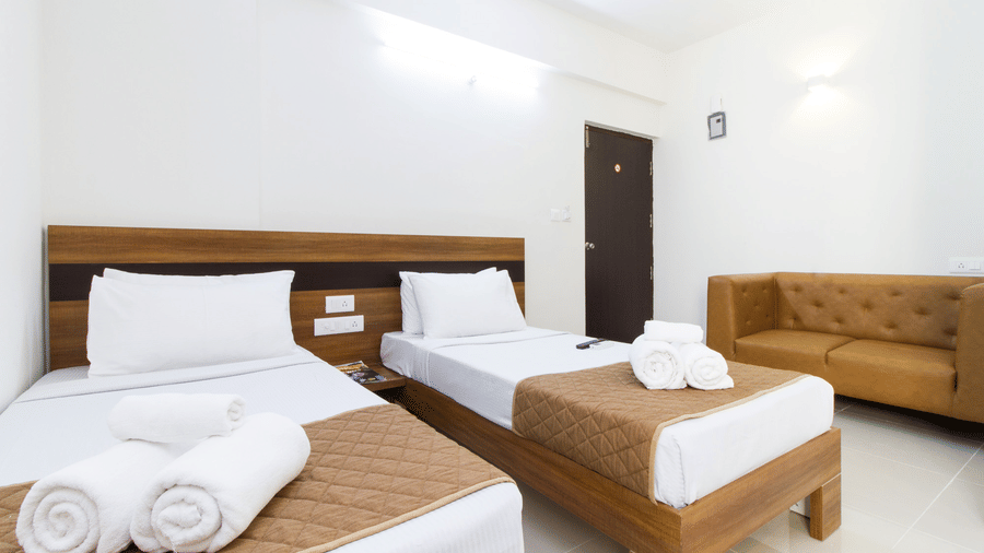 Twin bedroom with two beds, white linen, a sofa, and a wooden door at Sanctum Suites Whitefield, Bangalore.