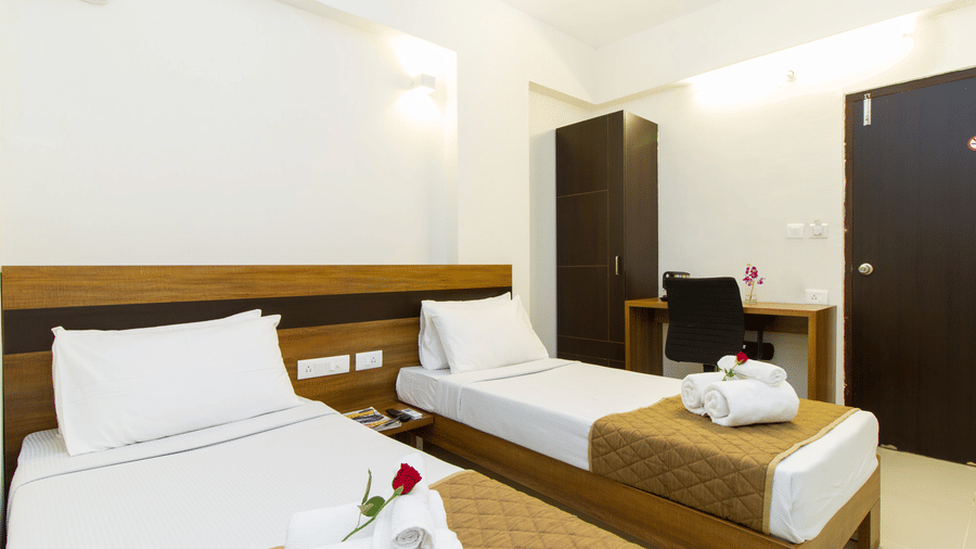 Twin bedroom with two beds, white linen, a dark wooden wardrobe, and a red rose accent at Sanctum Suites Whitefield, Bangalore.