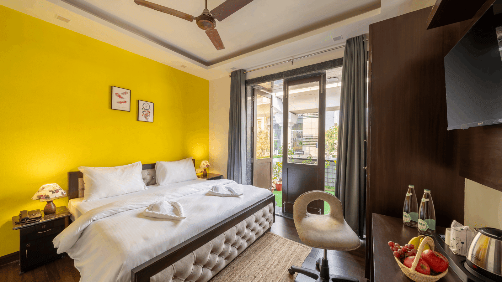 An image of a bedroom featuring a double size bed, a chair, a flat TV screen, a table with fruit basket and tea and coffee maker, and  a glass door leading to the balcony - Lime Boutique Bed & Breakfast, Nehru Place Metro
