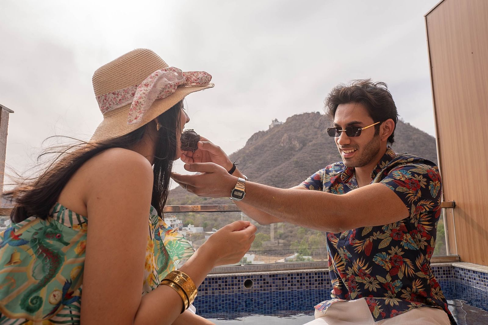 Two people sitting outdoors. The person on the left is wearing a hat and has long dark hair. The person on the right is wearing sunglasses and a patterned shirt.