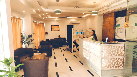 A view of the reception area at Hotel Apple Park Delhi, showing a staff member behind a backlit decorative desk and a seating area with brown leather sofas.