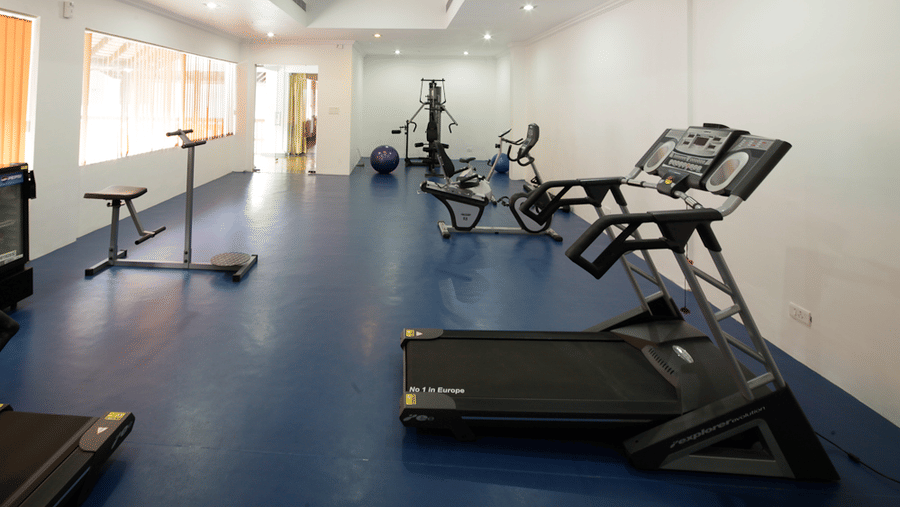 A fitness centre with white walls and a blue floor equipped with a treadmill, exercise bikes, and other gym machines | Le Pondy