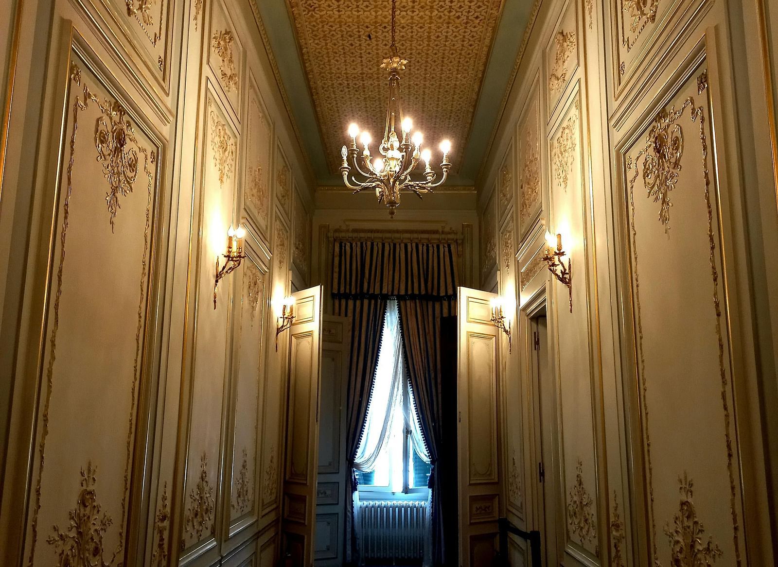 A dimly lit narrow hallway with white walls and European patterns on the wall  and there is a window at the end of the hallway