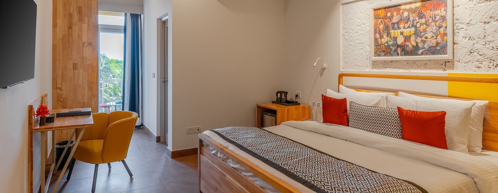 Bright hotel room with, flat TV screen, work desk and chair, vibrant art on the wall, and an access to the balcony at Bedzzz Xclusiv Morjim, Goa