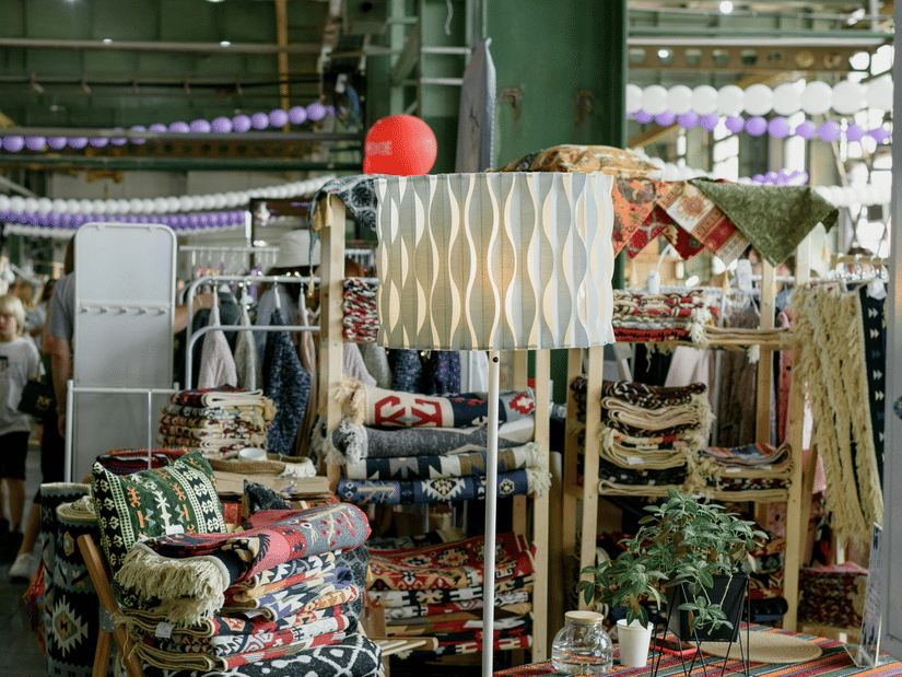 A busy indoor market stall featuring colourful textiles, a tall floor lamp, and purple bunting hung from the ceiling.