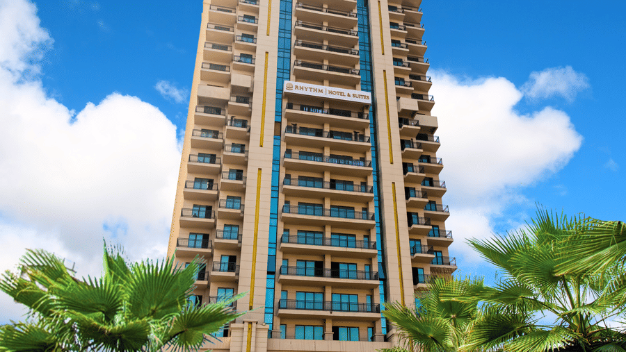 Front facade of Rhythm Gurugram surrounded by palm trees and landscaped greenery.