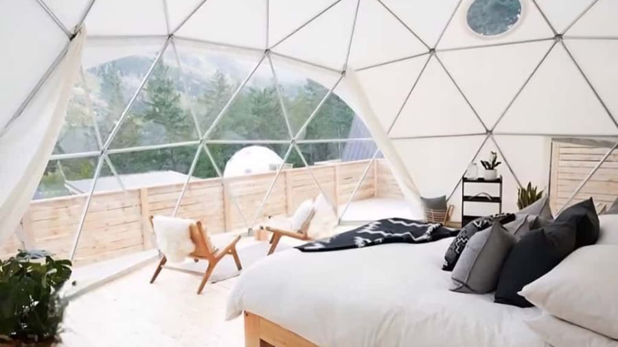 Interior of a Glass Dome room featuring a cozy bed, wooden flooring, and large windows offering a scenic view of nature.