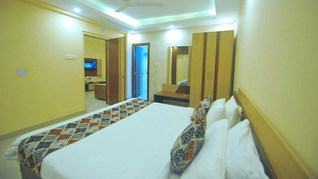 A well-lit bedroom at Pipul Hotel And Resort, Puri, featuring a bed, a ceiling fan, wardrobe, and an adjacent living area with a television.
