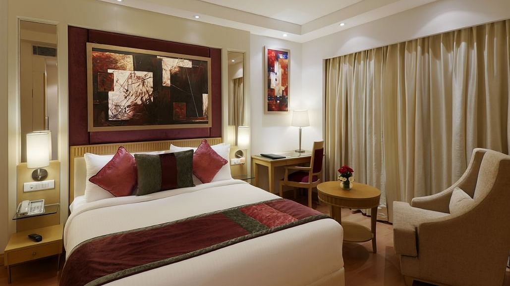 A beautifully designed bedroom featuring a double bed, a sofa chair with a small coffee table, and wall decor such as paintings – Inde Hotel Vista Woods, Gurgaon.