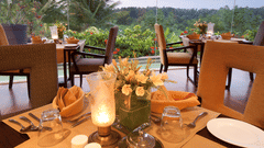 Candle-lit dining tables with plates, cutlery, napkins and floral centrepieces placed indoors, facing large glass windows that overlook greenery and palm trees at The Serai Chikmagalur.