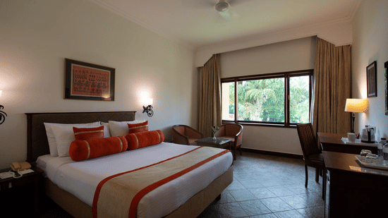 The Deluxe King Garden View bedroom at Clarks Khajuraho featuring a large bed with an upholstered headboard, side tables with lamps, and a work desk placed near a window with curtains allowing natural light.