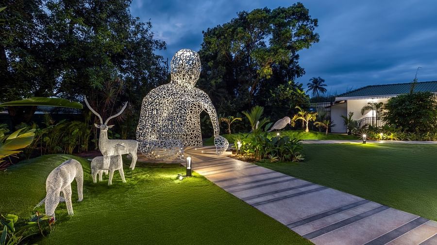  Illuminated outdoor sculptures of a human figure and deer on a well-manicured lawn, surrounded by tropical plants at dusk - Heritage Village Resorts & Spa, Goa