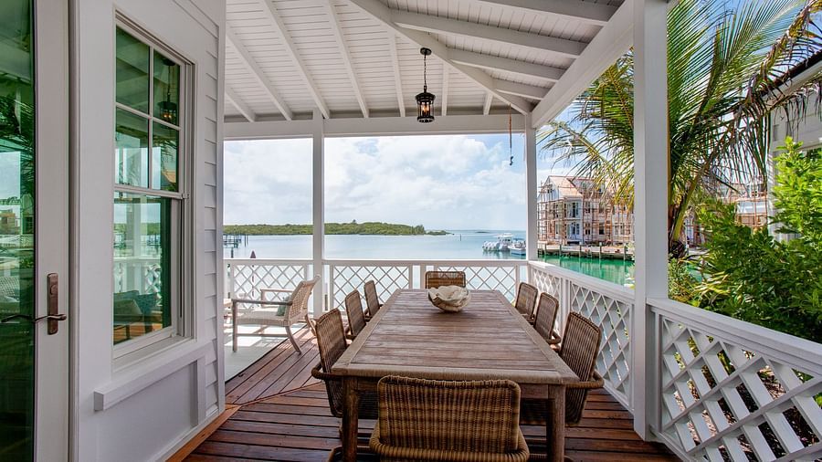 An outdoor dining area on a covered porch, featuring a long wooden table and chairs, with a view of a body of water and distant buildings - Abaco Inn