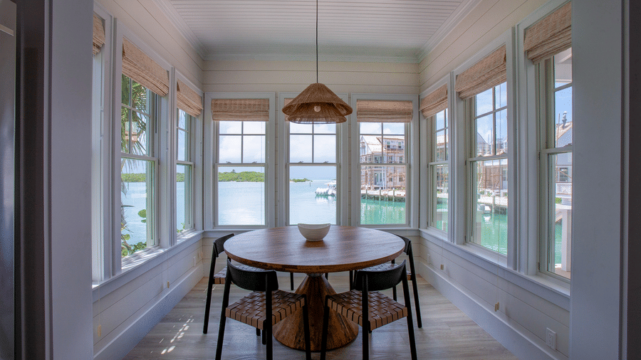 A bright dining area of Shoal Time Villa at Abaco Inn with a wooden table, chairs, large windows on all sides, and a hanging light fixture.