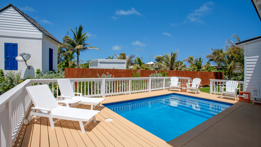An outdoor pool area at Shoal Villa in Abaco Inn with a rectangular swimming pool, wooden deck, sun loungers, surrounding buildings, and palm trees.