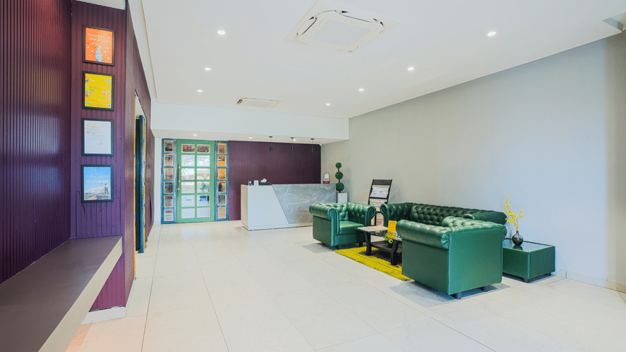 A bright, minimalist hotel lobby with a white reception desk, green leather sofas, and small framed art on a purple feature wall at Apartel by Aarin, Oragadam.