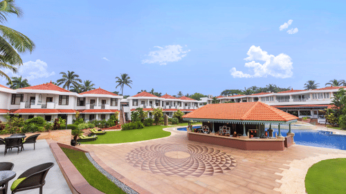 A stunning view of a resort with white-walled villas, red-tiled roofs, a landscaped garden, and a poolside bar under a bright blue sky - Heritage Village Resorts & Spa, Goa